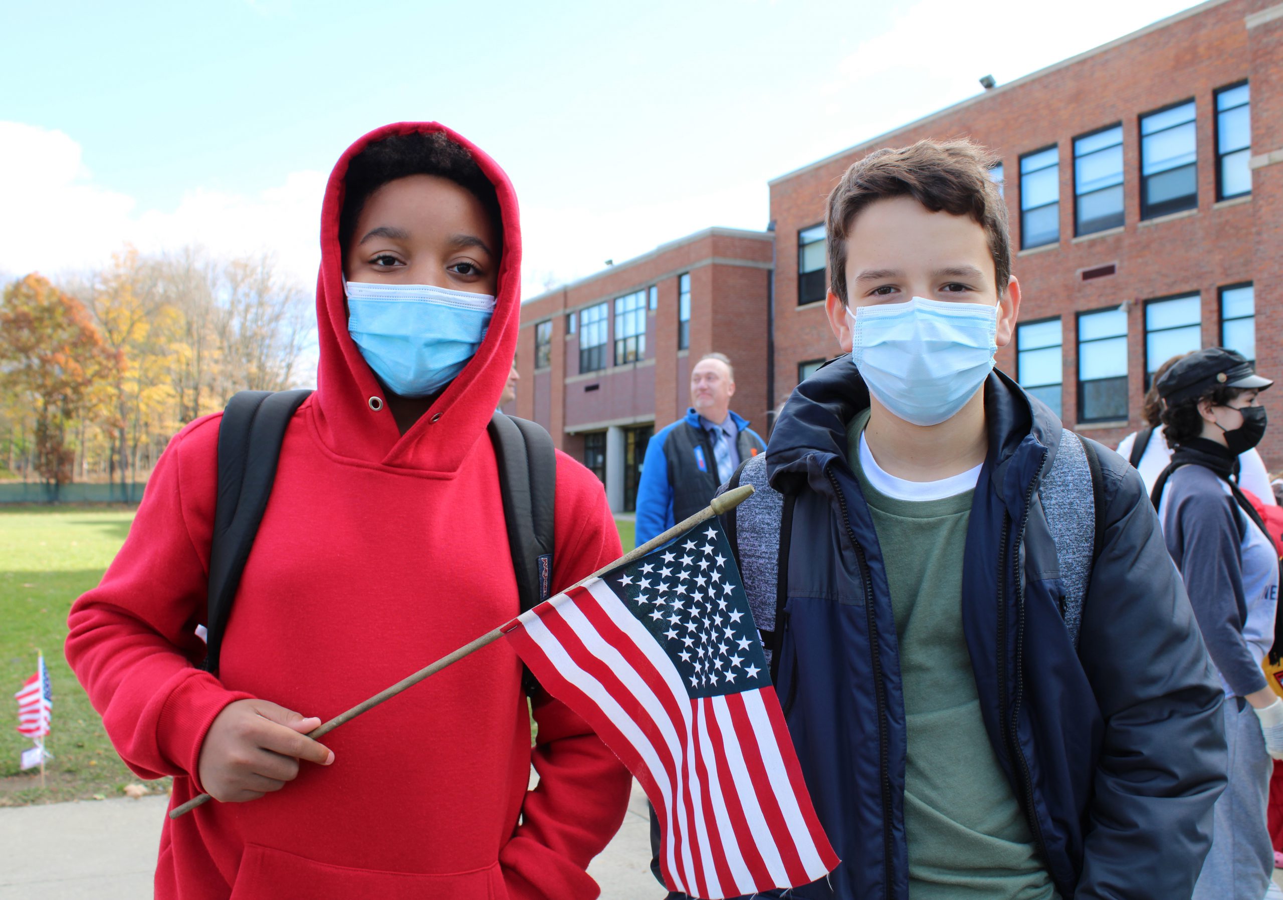 Two middle school boys stand outside, a brick building behind them and a blue sky. On the left the boy is wearing a red hooded sweatshirt and blue mask. He is holding an American flag. The boy on the right is wearing a blue jacket, green shirt and a blue mask.
