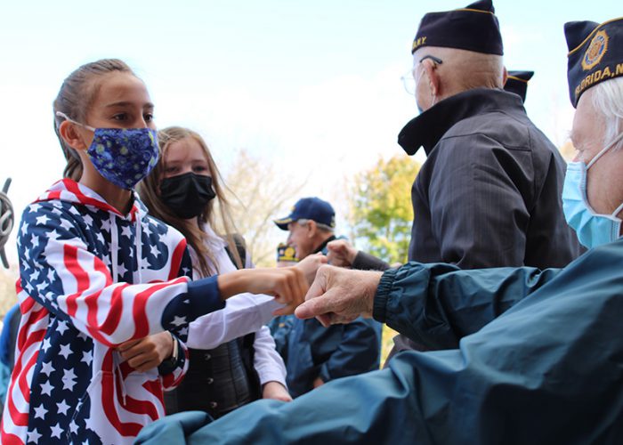 A middle school girl with long brown hair pulled back in a ponytail is wearing a sweatshirt that looks like an American Flag. She has a blue printed mask on and is fist bumping an older man who is seated, wearing a mask and a veterans group hat.