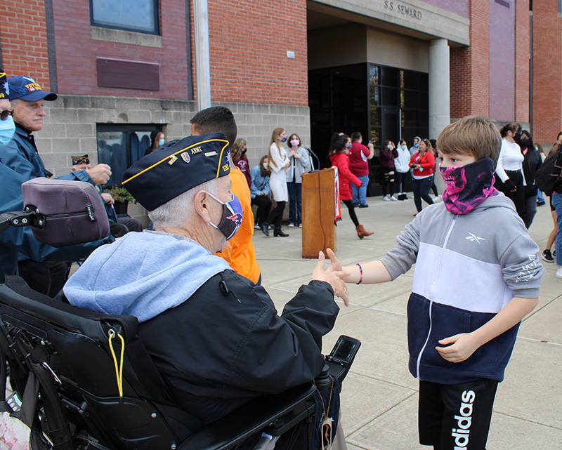 A middle school boy wearing a blue, white and gray hoodie and a blue mask shakes hands with an older man in a wheelchair, wearing a cap signifying he was a member of the army. He is wearing a facemask as well.
