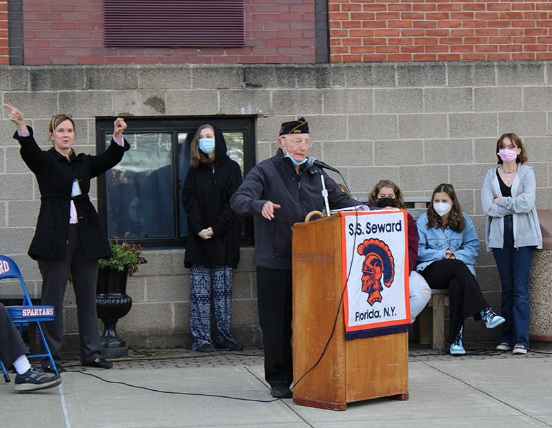 An older man, wearing navy blue jacket and a cap from a veterans organization, stands at a wooden podium with an S.S. Seward banner on it. Behind him are high school girls listening and a woman with her arms up cheering.