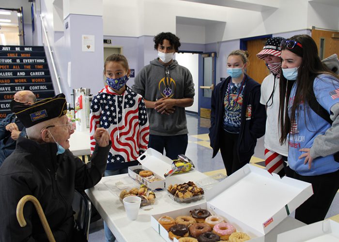 A man is seated. He is holding a cane at his side and has a cap on signifying he is a veteran. In front of him are donuts and cookies on a table. There five high school students standing around him listening to him talk, all looking at him directly. The students are wearing masks.