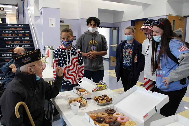 A man is seated. He is holding a cane at his side and has a cap on signifying he is a veteran. In front of him are donuts and cookies on a table. There five high school students standing around him listening to him talk, all looking at him directly. The students are wearing masks.