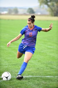 A girl in a blue and orange soccer uniform, number 12. She is in the process of kicking a ball.
