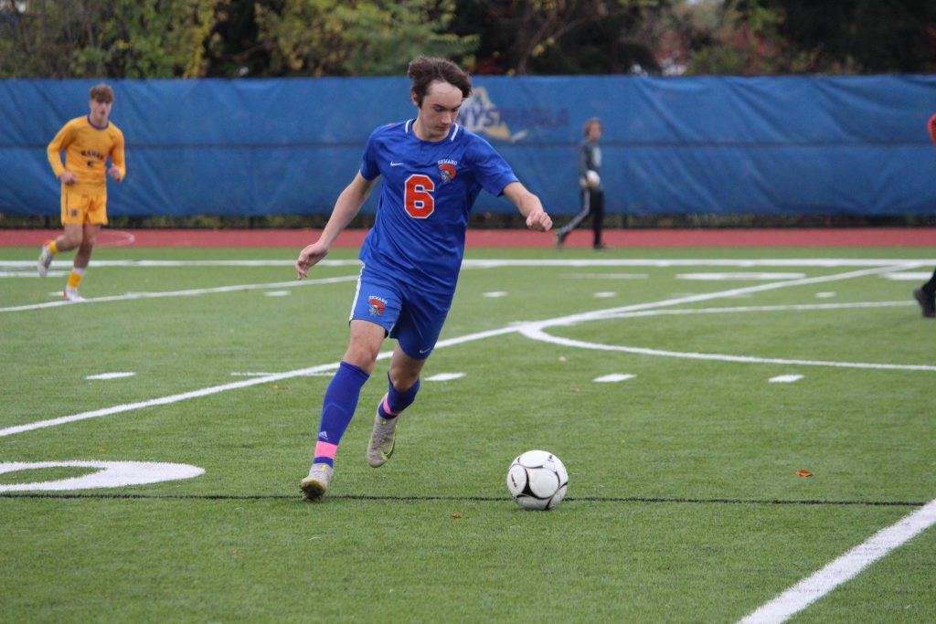 A high school boy dressed in a blue soccer uniform with the number  6 in orange. He is on a field and about to kick the ball.