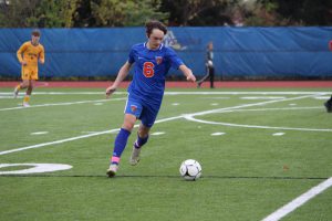 A high school boy dressed in a blue soccer uniform with the number 6 in orange. He is on a field and about to kick the ball.