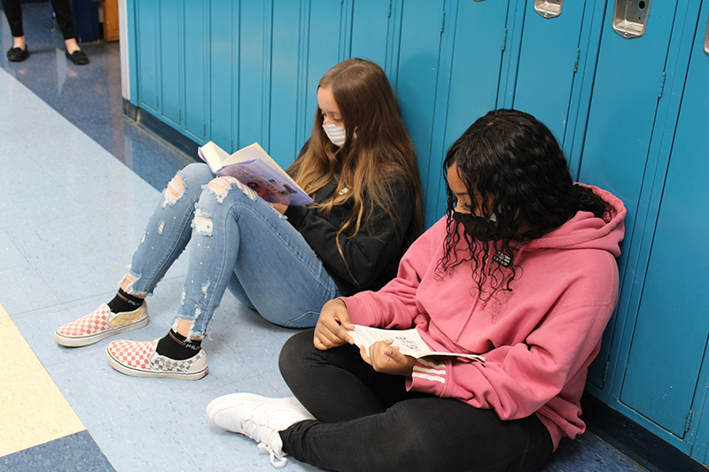Two middle school girls sit on a floor with blue lockers behind them. They are both reading books.