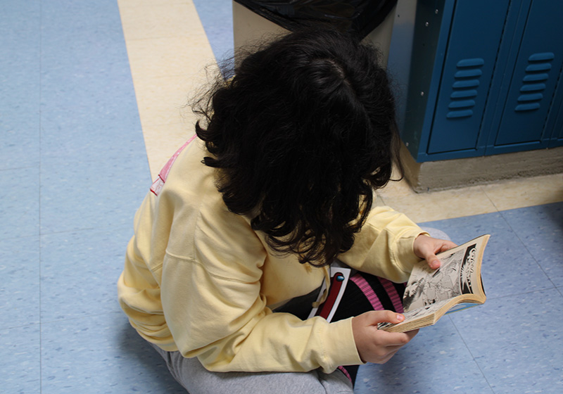 A middle school student sits on the floor with a book open.