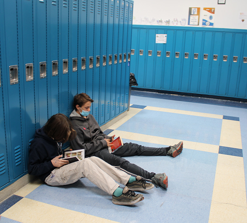 Two middle school boys are sitting on a floor in a hallway, leaning against blue lockers. They are both reading books.
