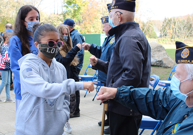 A line of high school students walk past a line of older men who are veterans. They are fist bumping them as they pass. All have masks on.