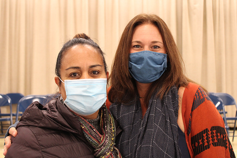 Two women stand together with their arm around each other. The woman on the left has her hair pulled back and is wearin g alight blue mask, brown jacket and scarf. The woman on the right has long brown hair and is wearing a blue mask, orange and black sweater.
