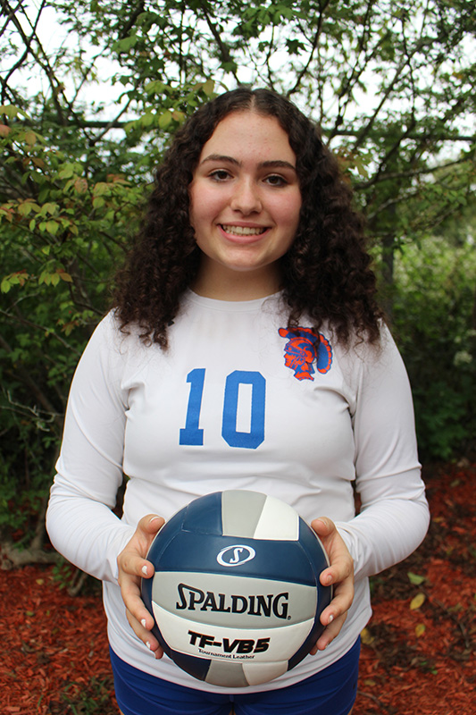 A high school girl with long dark hair holds a volleyball and smiles. She is wearing a white long-sleeved volleyball shirt with the number 10.
