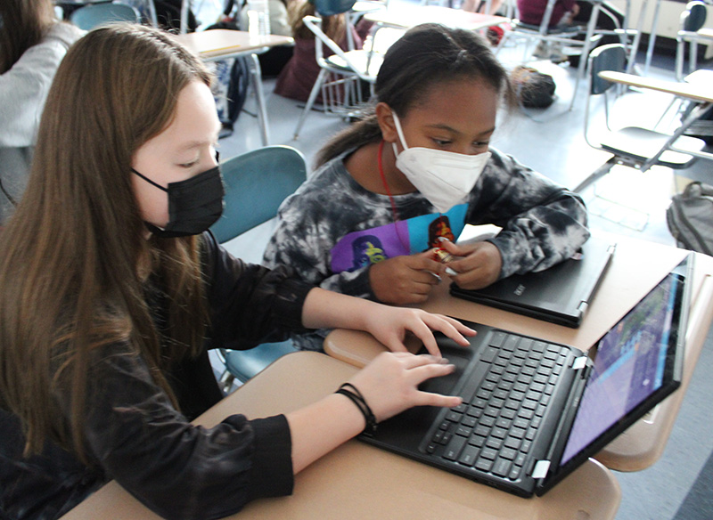 Two girls look at a chromebook. The one on the left has long brown hair and is wearing a dark shirt and mask. Her hands are on the chromebook typing. The girl on the right has her long dark hair pulled back, is wearing a white mask and a printed shirt.