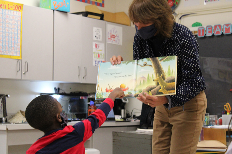 A woman in a blue blouse with white polka dots and tan pants and a dark mask, holds open a children's book. A second-grade boy, wearing a dark mask and a red and blue striped shirt, points to something on the page.