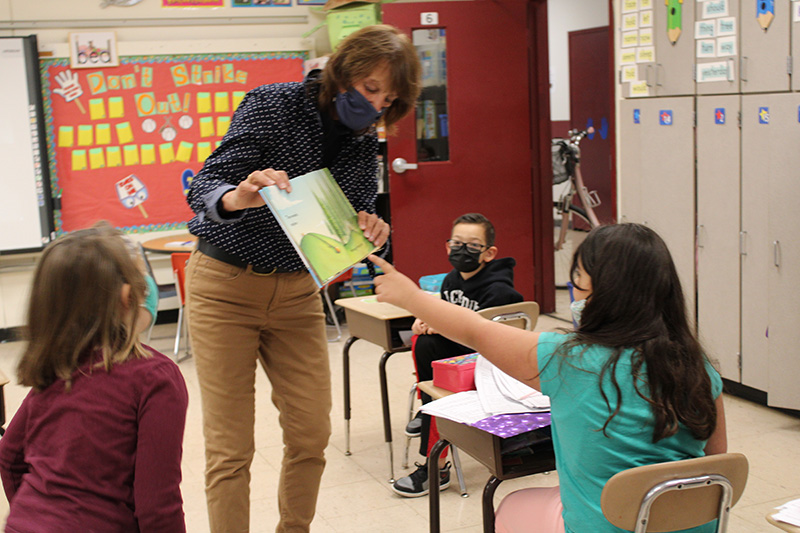 A woman in a blue blouse with white polka dots and tan pants wearing a dark mask, holds open a book. A girl with long dark hair wearing a blue green shirt points to something on the page. Two other students watch.