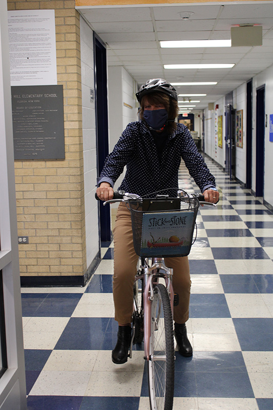 A woman wearing a blue blouse with white polka dots, tan pants, dark mask and a bicycle helmet rides a bike with a basket in front. In the basket is a book. The floor is white and blue squares.