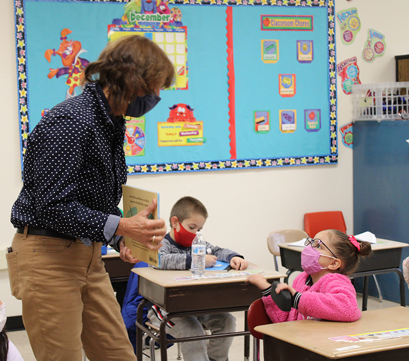 A woman wearing a blue blouse with white polka dots, tan pants, dark mask  holds a book open as two second-grade students watch. One of the students, a girl wearing a pink shirt with a pink bow in her hair and glasses looks up at the woman. She also has a pink mask on.