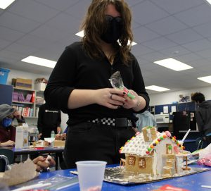 A high school girl, dressed in black with long dark hair, decorates an almost completed gingerbread house. It is very colorful.