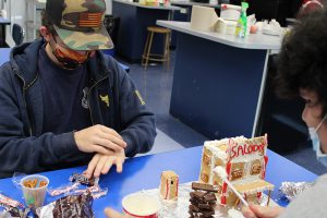 Two high school boys decorate a gingerbread house. It is almost complete and has SALOON written in candy above it.