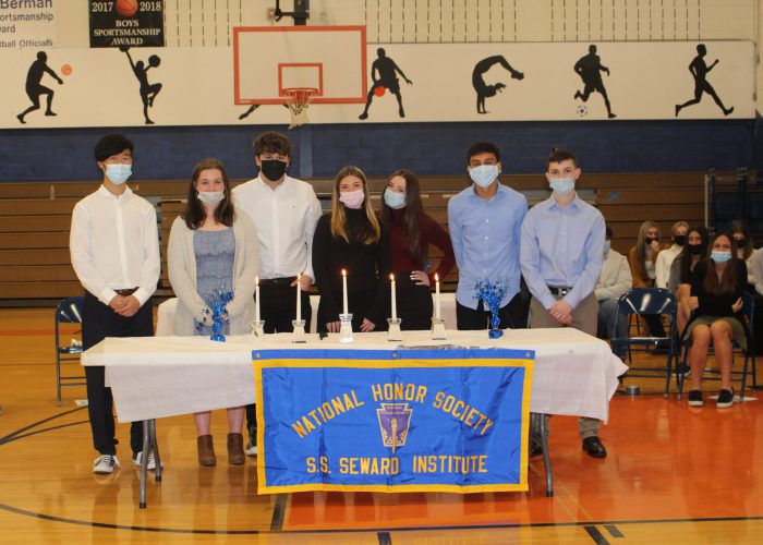 A group of seven high school students standing around a table. Three girls and four boys. They are all wearing masks. On the table are four long candles. There is a banner on the front of the table that says National Honor Society S. S. Seward Institute. They are in a gym.