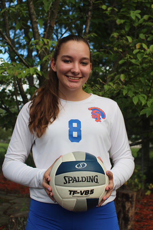 A high school girl with long dark hair holds a volleyball and smiles. She is wearing a white long-sleeved volleyball shirt with the number 8.