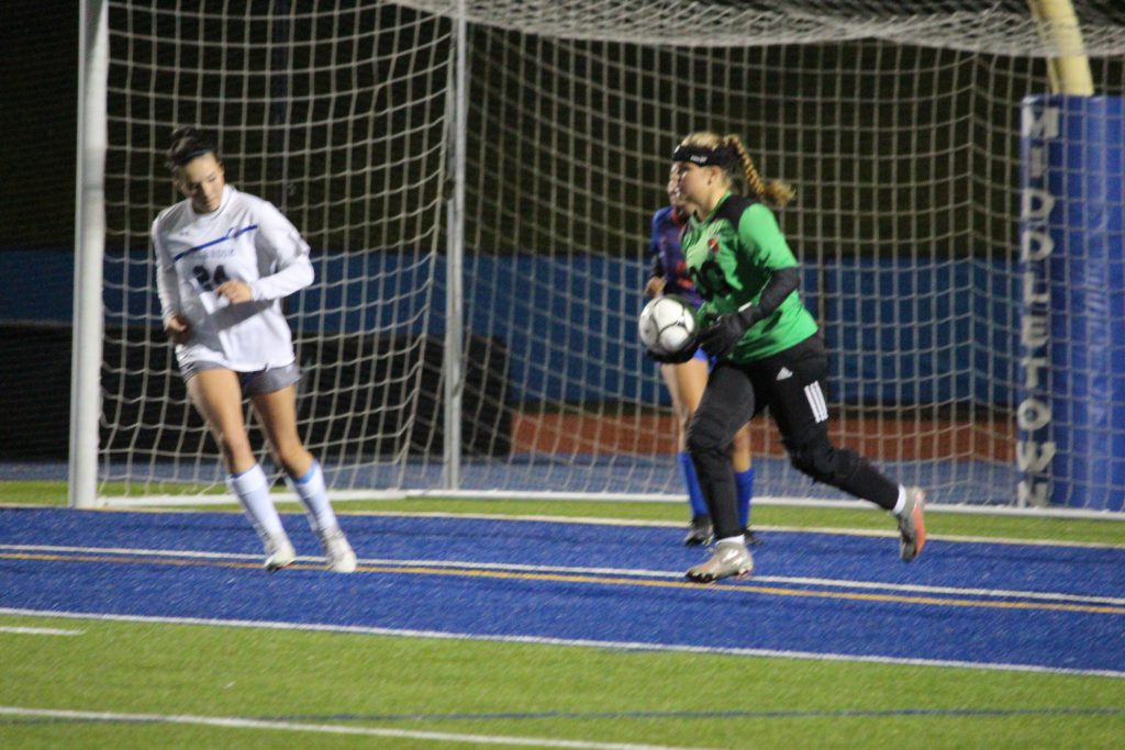 A high school girl in a green goalie's shirt and black pants kicking a soccer ball away.