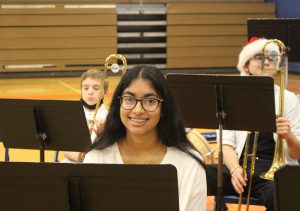 A middle school student with long dark hair and glasses, wearing a white shirt, smiles from behind a music stand. There are two students behind her, holding instruments.