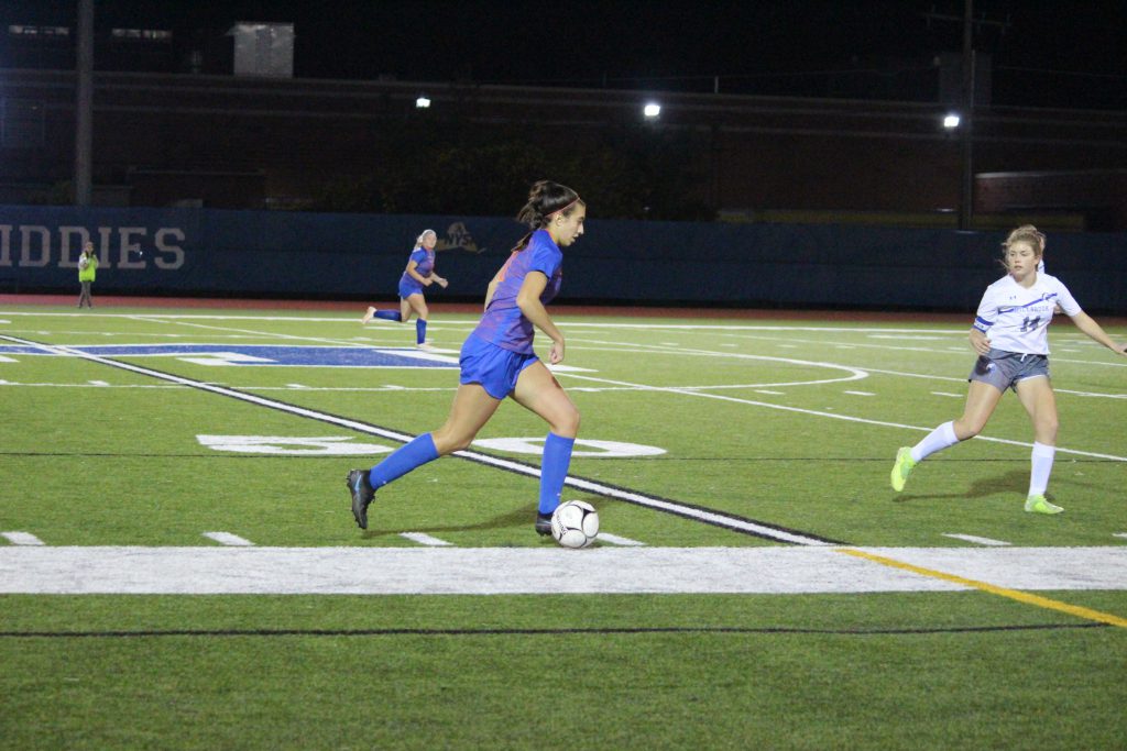A high school soccer player wearing a blue soccer uniform dribbles the ball along the sideline. She has dark hair pulled back in a ponytail.