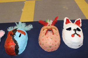 A table with a dark blue table cloth and three handmade masks on it. One is a white dog, the one in the center looks like a chicken and the one on the left is half blue and half red and black.