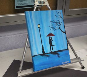 A painting on an easel. It is mostly a blue background with a streetlamp. Under the streetlamp is a couple hugging each other holding a red umbrella.