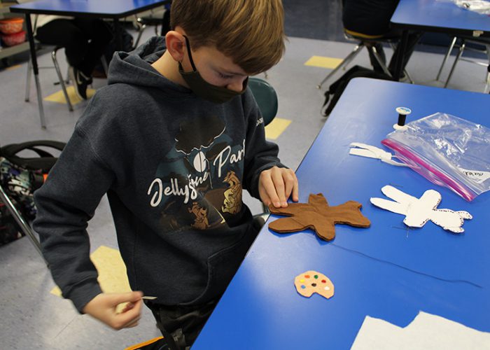 A younger middle school age young man, wearing a blackmask and gray sweatshirt, sews a small brown bear , which is on a blue table. There is a small palate next to the bear.