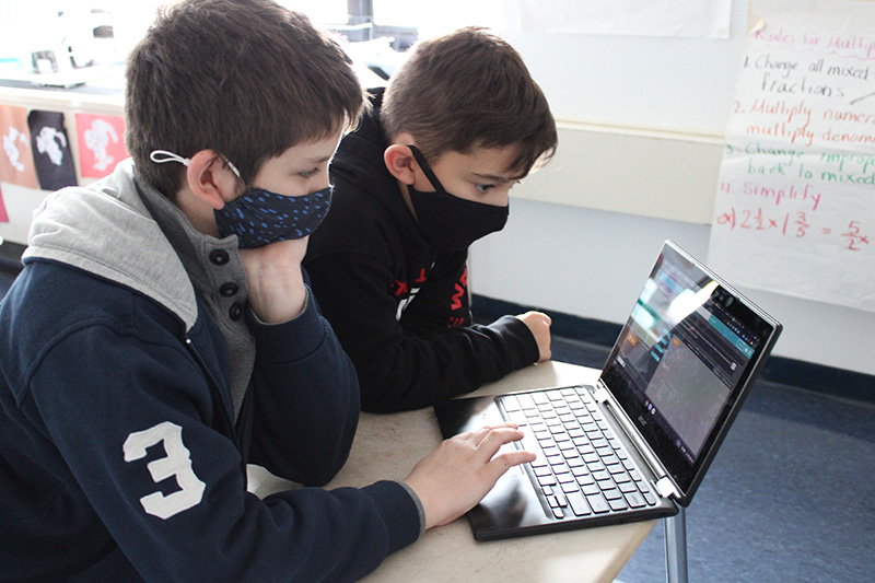 Two middle school boys, both wearing masks, look at a chromebook. One has his hand on the keyboard. They are looking intensely.