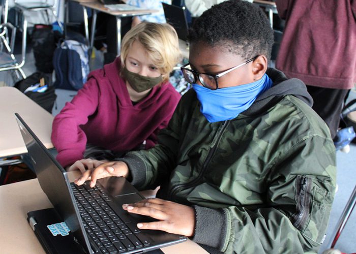 Two boys, both wearing masks, one wearing a red sweatshirt, the other with a green jacket, and glasses, look at a chromebook. The boy on the right is typing on the keyboard.