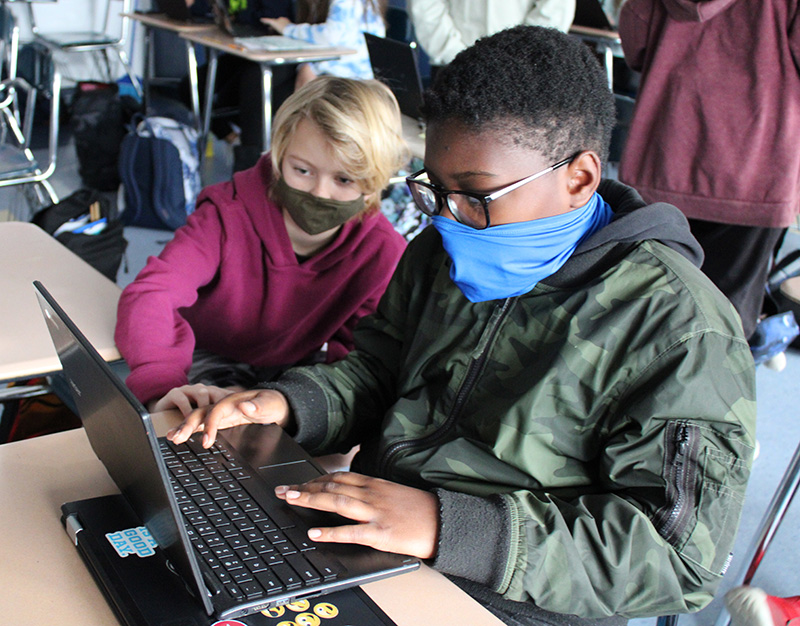 Two boys, both wearing masks, one wearing a red sweatshirt, the other with a green jacket, and glasses, look at a chromebook. The boy on the right is typing on the keyboard.