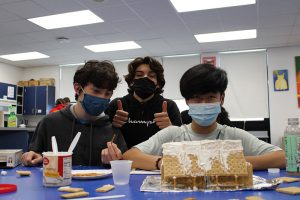 Three high school boys all wearing masks build a gingerbread house. The boy in the center is giving two thumbs up.