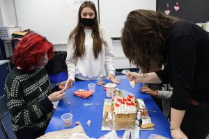 Three high school girls building a gingerbread house. The girl on the right is decorating it. All are wearing masks.
