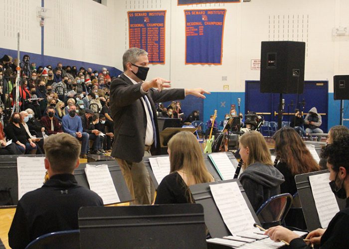 A man standing up with a jacket and tie on and black mask. He has his ars out wide ready to conduct a band of many high school students sitting in front of him with music on their music stands.