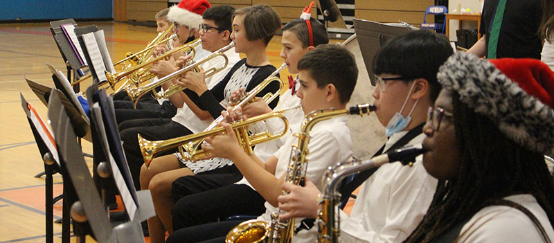 A line of seven students playing wind instruments - horns and saxophones. A couple of the students have festive hats and headbands on.