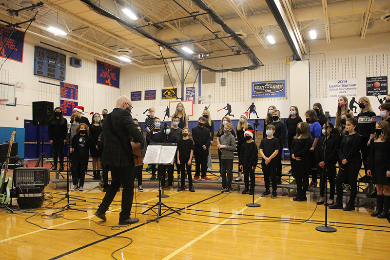 A man whose back is to the camer, plays guitar. In front of him is a group of students mostly dressed in black singing. They are in a large gymnasium.