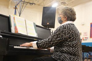 A woman with short dark hair, wearing glasses and a mask and a black and white sweater, plays a black piano.