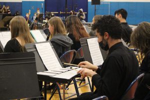 A young man dressed in black and a black mask, holds two musical hammers over a glockenspiel. There are other musicians around him.