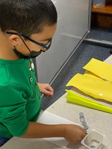 A younger elementary student with shor dark hair, glasses and a black mask and green shirt stands at a table counting coins.