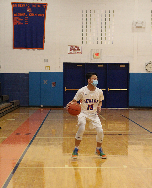 A shiny wooden floor in a gym. There is a young man wearing a white basketball uniform with the number 15. The jersey says Seward. He is wearing a blue mask and dribbling a basketball.