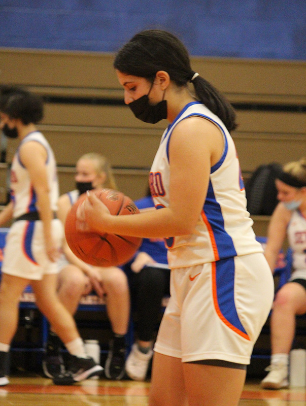 A photo from the side, a young woman wearing a white basketball uniform, with blue and orange stripes down the side. She has her dark hair pulled back into a pony tail and is wearing a black mask. She is holding a basketball.