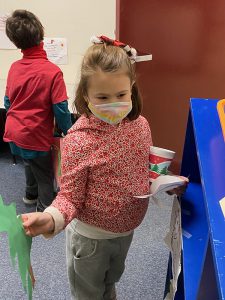 A second-grade girl wearing a pink patterned shirt and mask, holds a green tree made of construction paper.