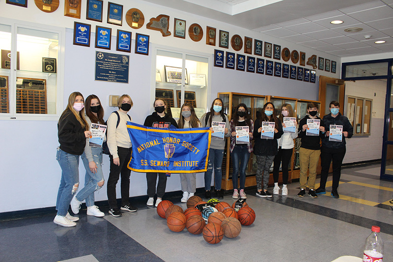 Eleven high school students lined up against a wall, They are all holding papers. Three toward the beginning of the line are holding a blue banner that reads National Honor Society on top and S.S. Seward Institute on the bottom in gold. In front are a bunch of basketballs on the floor.