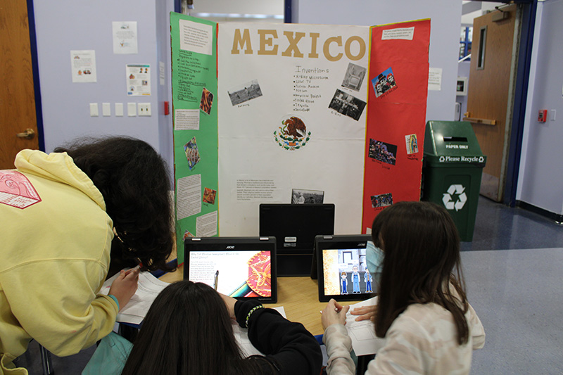 Three middle school students sit at a table writing while looking at Chromebooks. In front of them is a poster about Mexican immigration with the word Mexico on it.