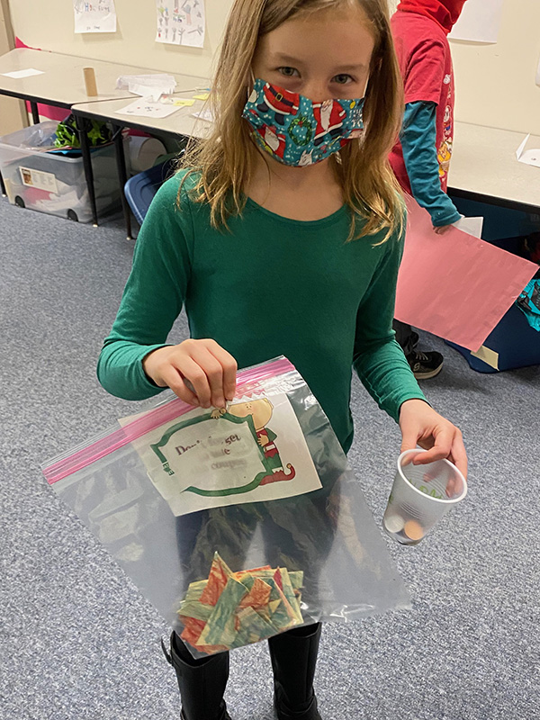 A young elementary girl wearing a green shirt and multi-color mask holds a bag of cut up paper and a cup with play coins.