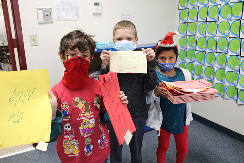 Three second-grade students stand holding paper products they created like a sleeping bag made from construction paper, a basket filled with different colored papers.