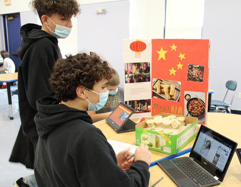 Two middle school boys, both with curly hair and masks and dark hoodies, look at a display with information about China immigration. One is sitting at a Chromebook. In the back are little containers of fried rice and the Chinese flag.
