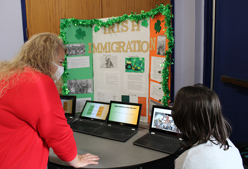 A woman with blond hair wearing a red shirt looks at a board about Irish immigration. A young woman sits to her right also watching.